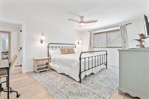 A bedroom with a bed, bedside tables, a desk, a chair, a ceiling fan, a window with curtains, and a dresser.