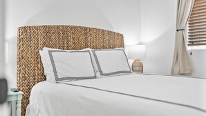 Una cama bien hecha con sábanas blancas y dos almohadas, un cabecero tejido, una mesita de noche con una lámpara y una ventana con cortinas.