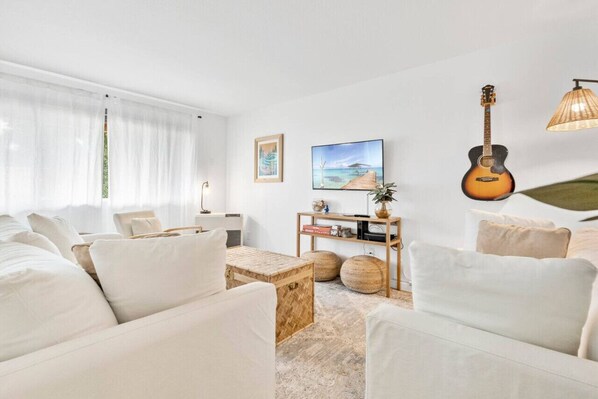 A modern living room with a white sofa set, a wooden coffee table, a guitar, and a TV on the wall.