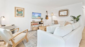 Una sala moderna con un sofá blanco, una silla de madera, una mesita con libros, una guitarra y decoración en la pared.
