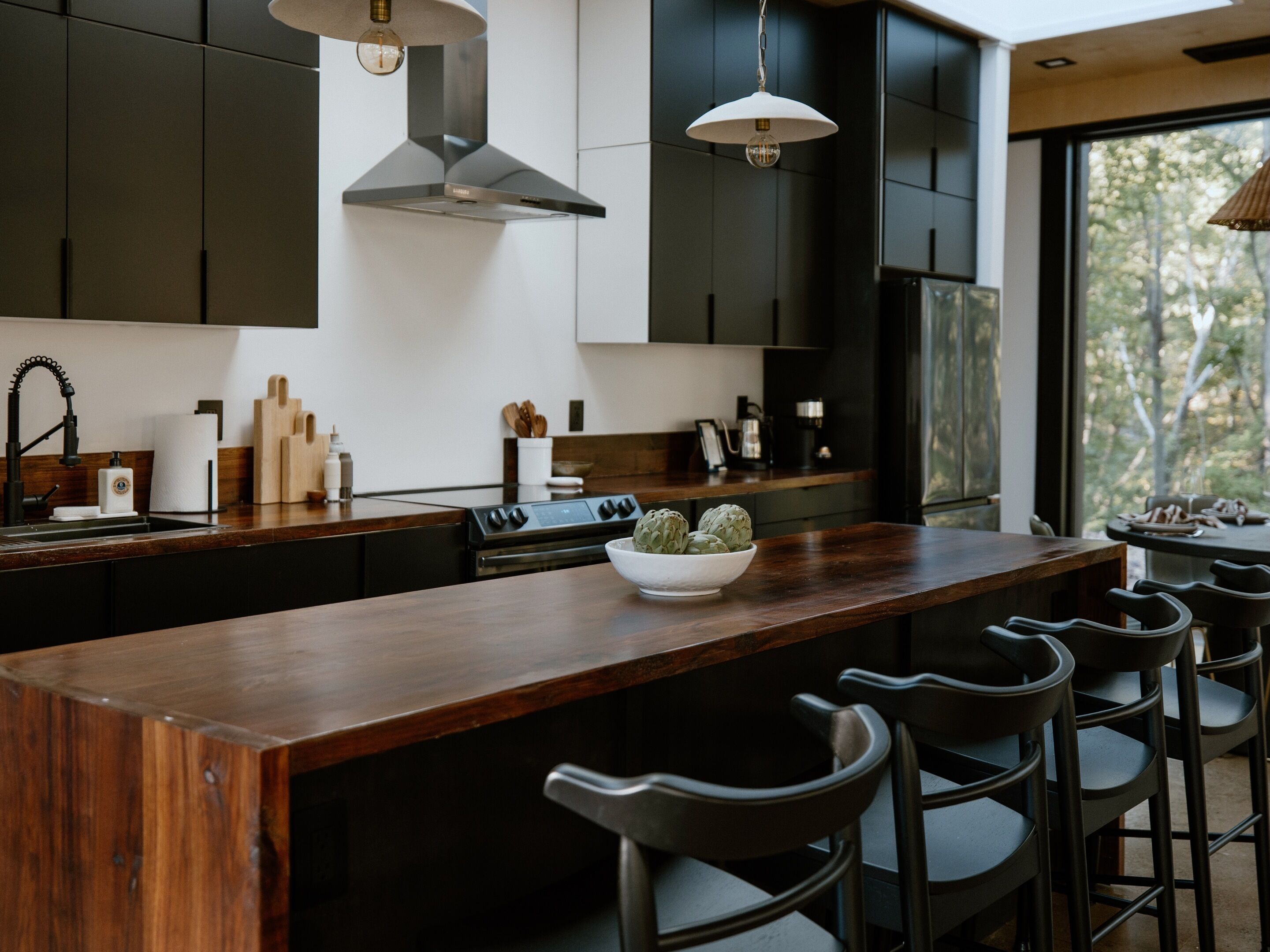 A modern kitchen with dark cabinetry, a wooden island, bar stools, and a large window offering a view of trees.