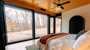 A bedroom with a large bed, a ceiling fan, and a view of a wooded area through large windows. - Ananda | Hocking Hills, Luxury 2BR Cabin, Hot Tub (Rockbridge)