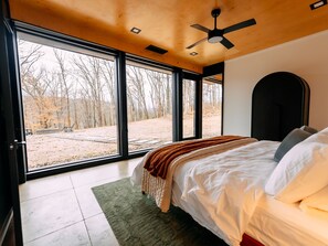 A bedroom with a large bed, a ceiling fan, and a view of a wooded area through large windows.