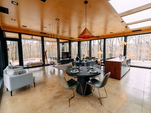 A modern dining area with a round table, chairs, and a large window offering a view of the forest.
