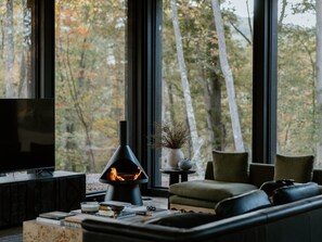 Ein modernes Wohnzimmer mit Kamin, Fernseher und Blick auf Bäume durch große Fenster.
