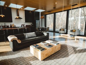 Ein modernes Wohnzimmer mit einer schwarzen Ledersofa, einem Couchtisch mit Büchern und einem großen Fenster mit Blick auf Bäume.