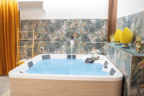 A hot tub in a room with tropical wallpaper, a wooden floor, and a yellow curtain.