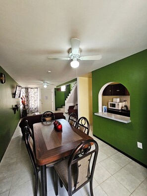 A dining area with a wooden table and chairs, a ceiling fan, and a kitchen area with a microwave and blender.
