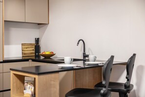 A modern kitchen with a black countertop, wooden cabinets, a black chair, a white sink, and a fruit bowl.