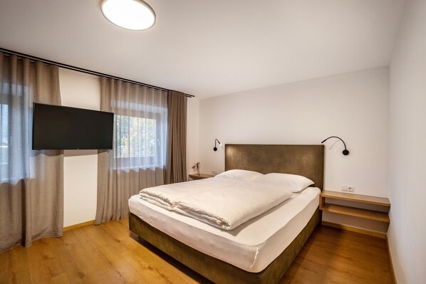 A bedroom with a bed, curtains, a TV, a bedside table, and a nightstand.