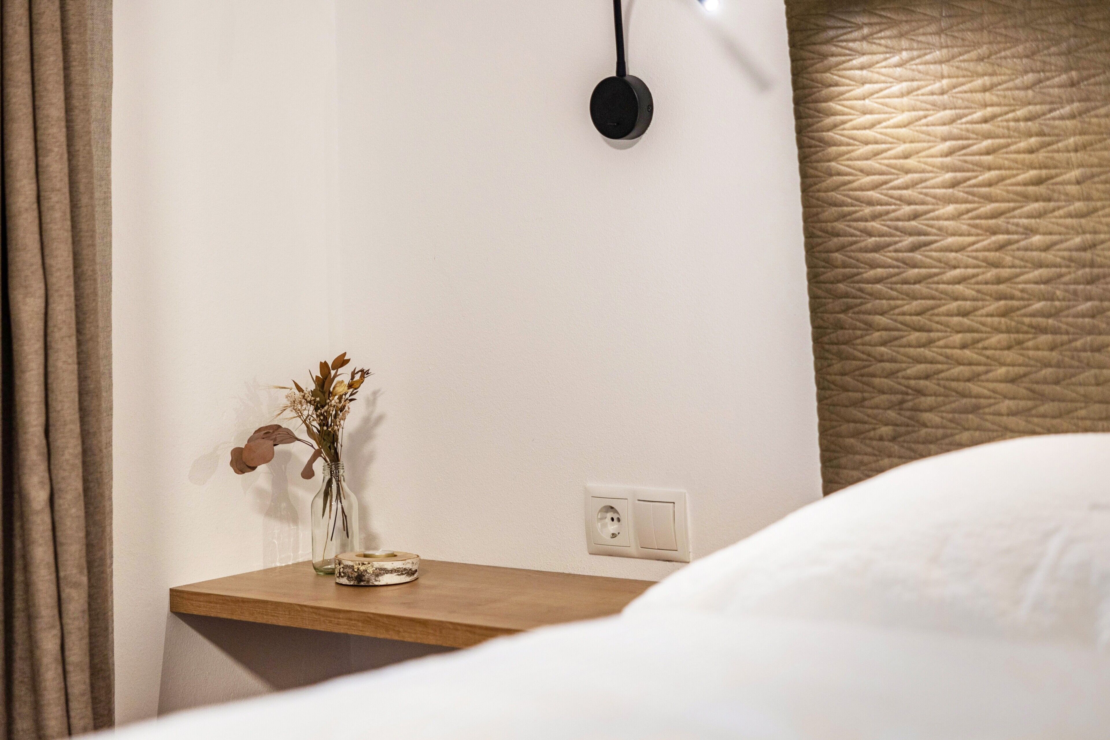 A hotel room with a wooden bedside table, a vase with dried flowers, a power outlet, and a textured wall.