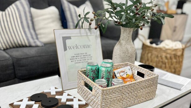 A modern living room with a coffee table featuring a welcome sign, a decorative plant, and a basket with snacks.
