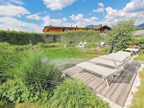 Une terrasse en bois avec des chaises longues, un petit étang et une maison en arrière-plan.