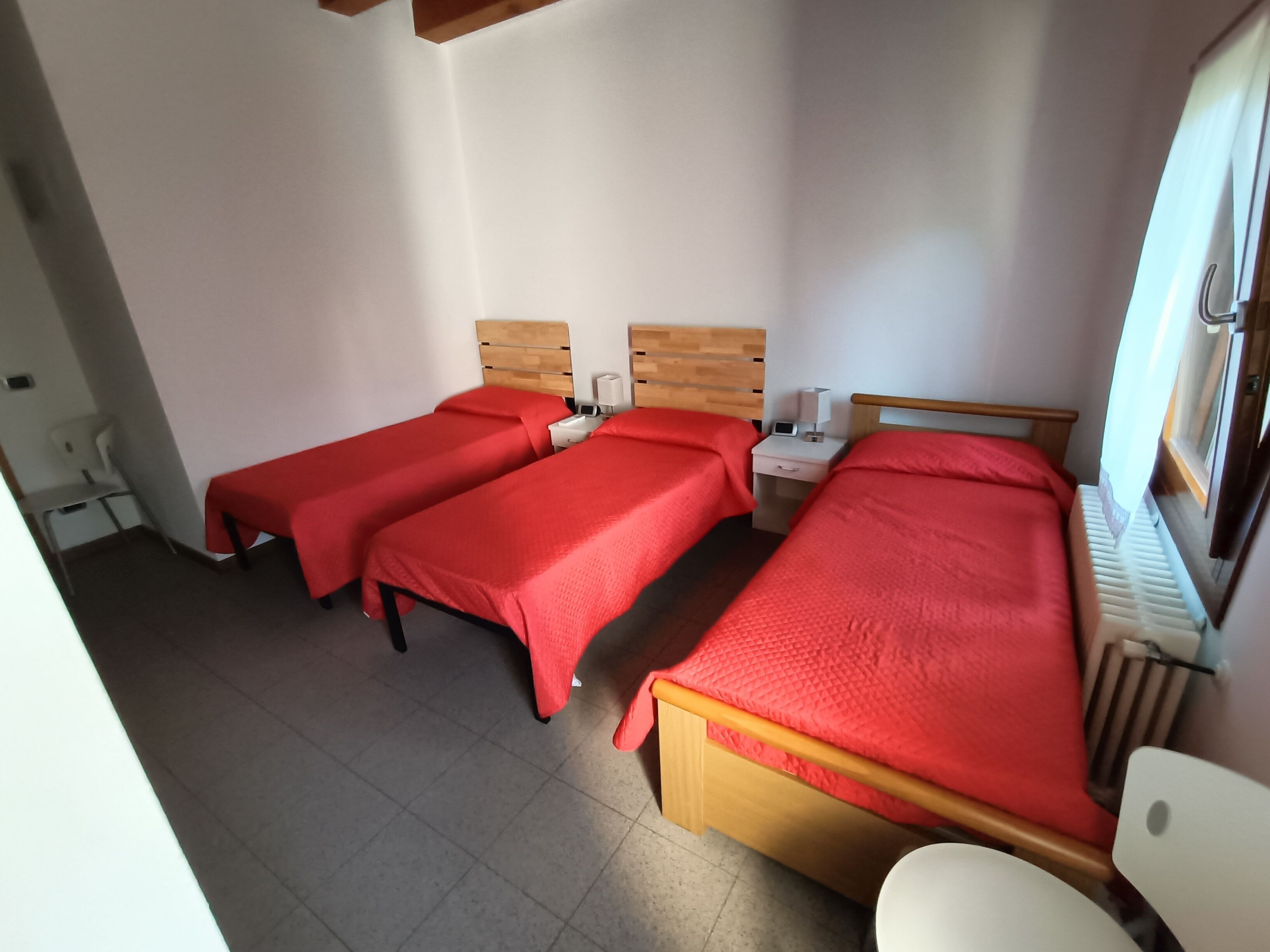 A room with three single beds, each with a wooden headboard and red bedding. There is a white bedside table with a lamp and a small drawer. The room has a tiled floor and a window with curtains.