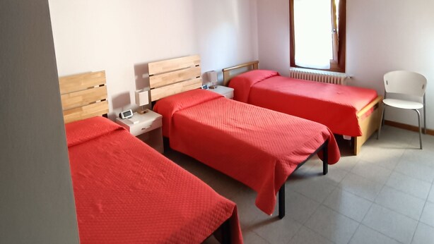 Una habitación con dos camas, cada una con colcha roja, cabeceros de madera y mesitas de noche blancas. Se ve una ventana con vistas al exterior, un radiador y una silla blanca.