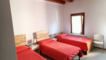 A room with two beds, each with a red bedspread, wooden headboards, and white nightstands. There is a window with a view of the outside, a radiator, and a white chair.
