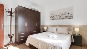 A bedroom with a large bed, a wardrobe, a coat rack, a bedside table with a lamp, and a wall-mounted artwork.