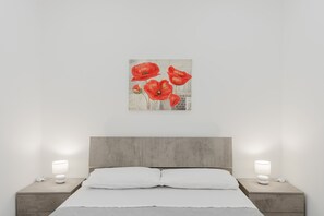 Un dormitorio con una cama, dos mesitas de noche con lámparas y un cuadro colgado en la pared de flores rojas.