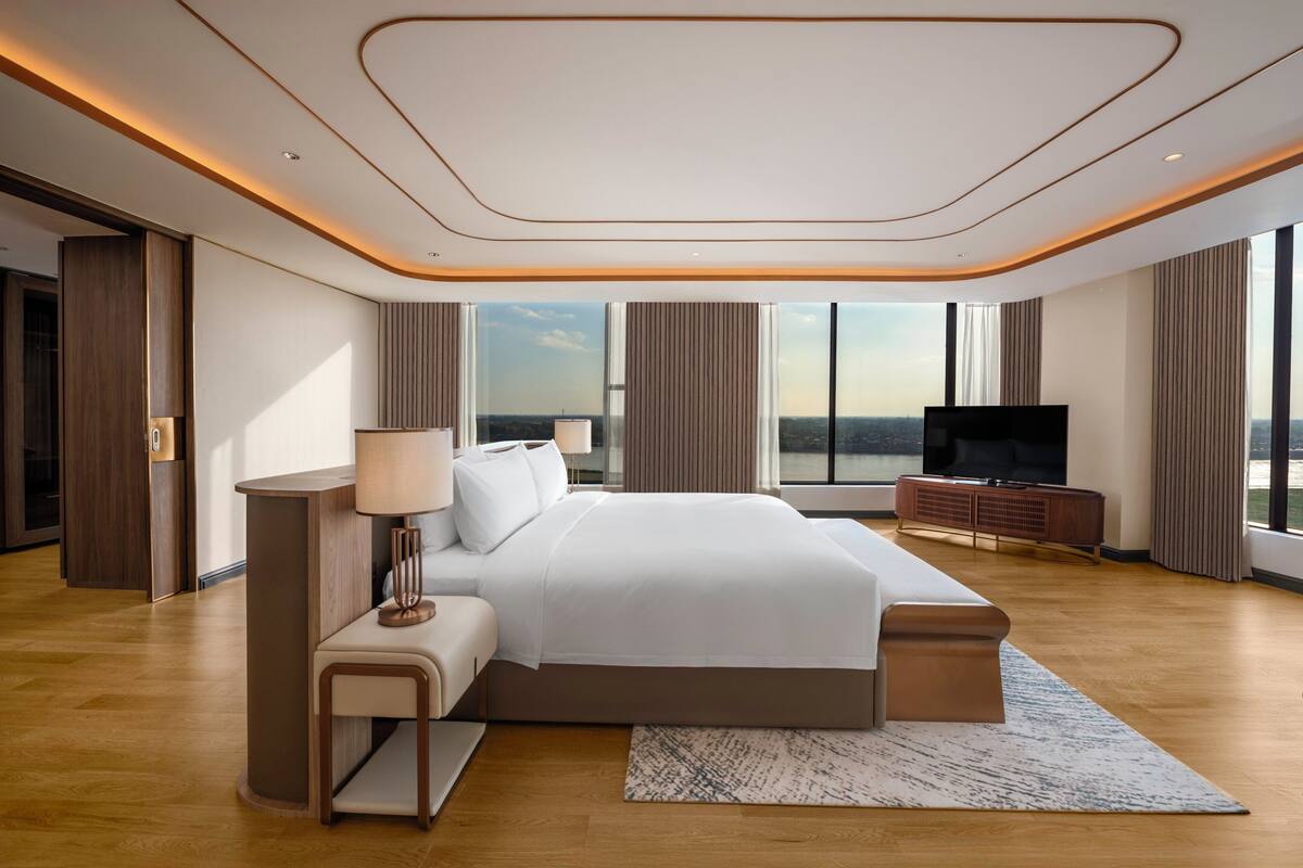 A modern hotel room with a large bed, a flat-screen TV, a wooden floor, and a view of the city.
