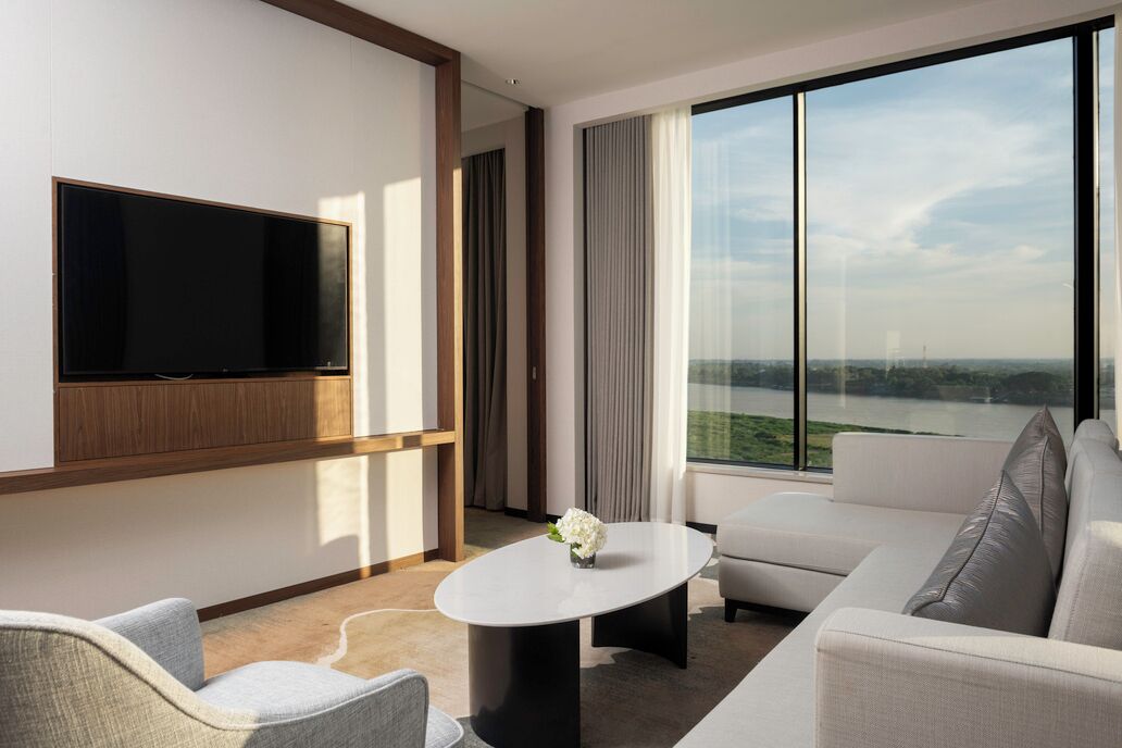 Une chambre d’hôtel moderne dotée d’un grand téléviseur à écran plat fixé au mur, d’un canapé confortable, d’une petite table ronde agrémentée d’un vase et d’une grande fenêtre offrant une vue sur une étendue d’eau et un paysage lointain.
