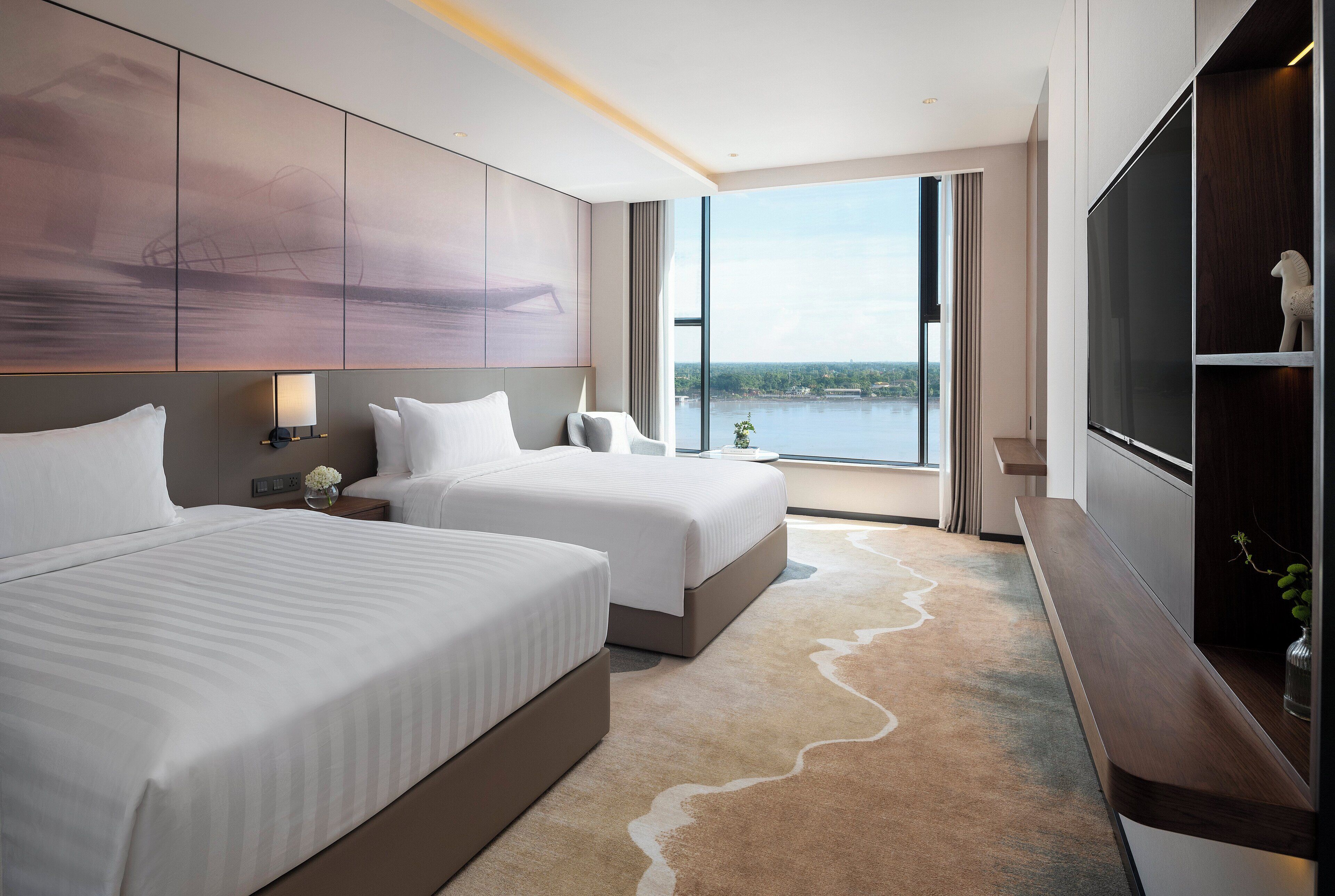 A modern hotel room with two beds, a large window offering a view of a body of water, and a flat-screen TV on a wooden console.