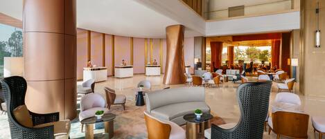 A spacious lobby with a modern design, featuring a central seating area with sofas and chairs, a reception desk, and a large pillar.