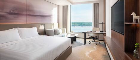 A modern hotel room with a large bed, a desk, a chair, a TV, and a view of the water.