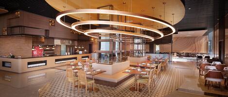 Um restaurante moderno com área de estar central, cozinha ao fundo e um grande lustre circular.
