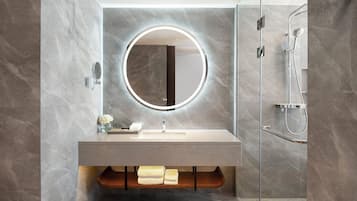 A modern bathroom with a marble wall, a round mirror with a light, a sink, and a glass-enclosed shower.