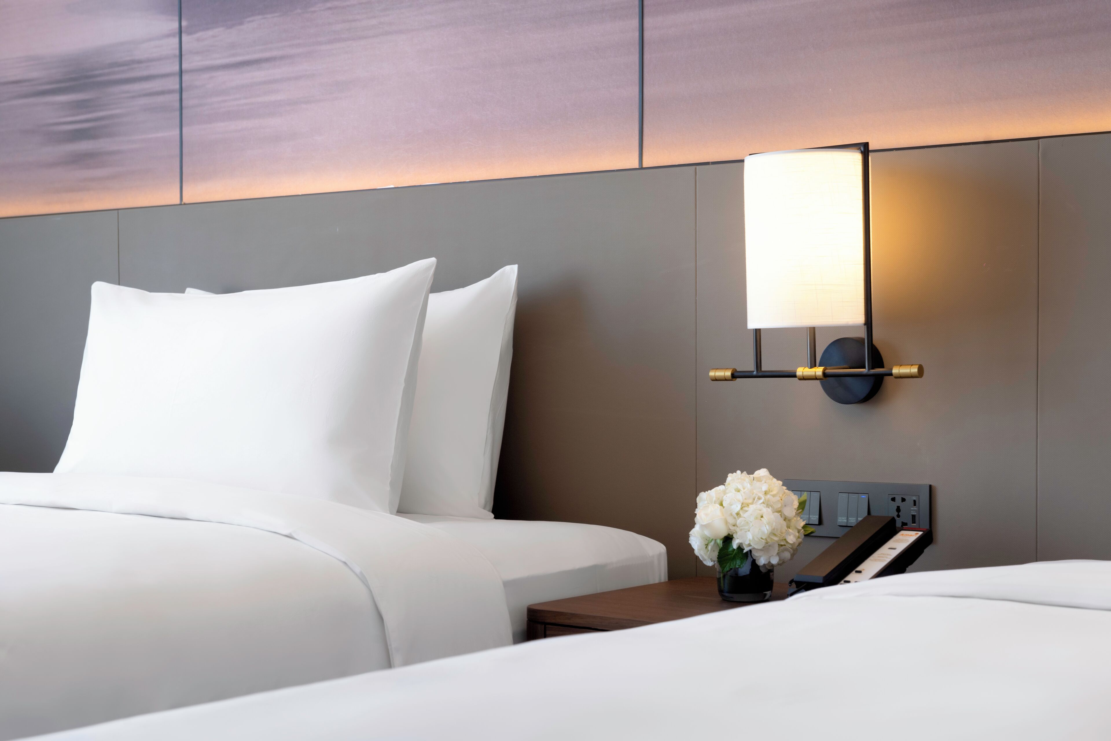 Una habitación de hotel con cama, mesita de noche, lámpara y panel de control en la pared.
