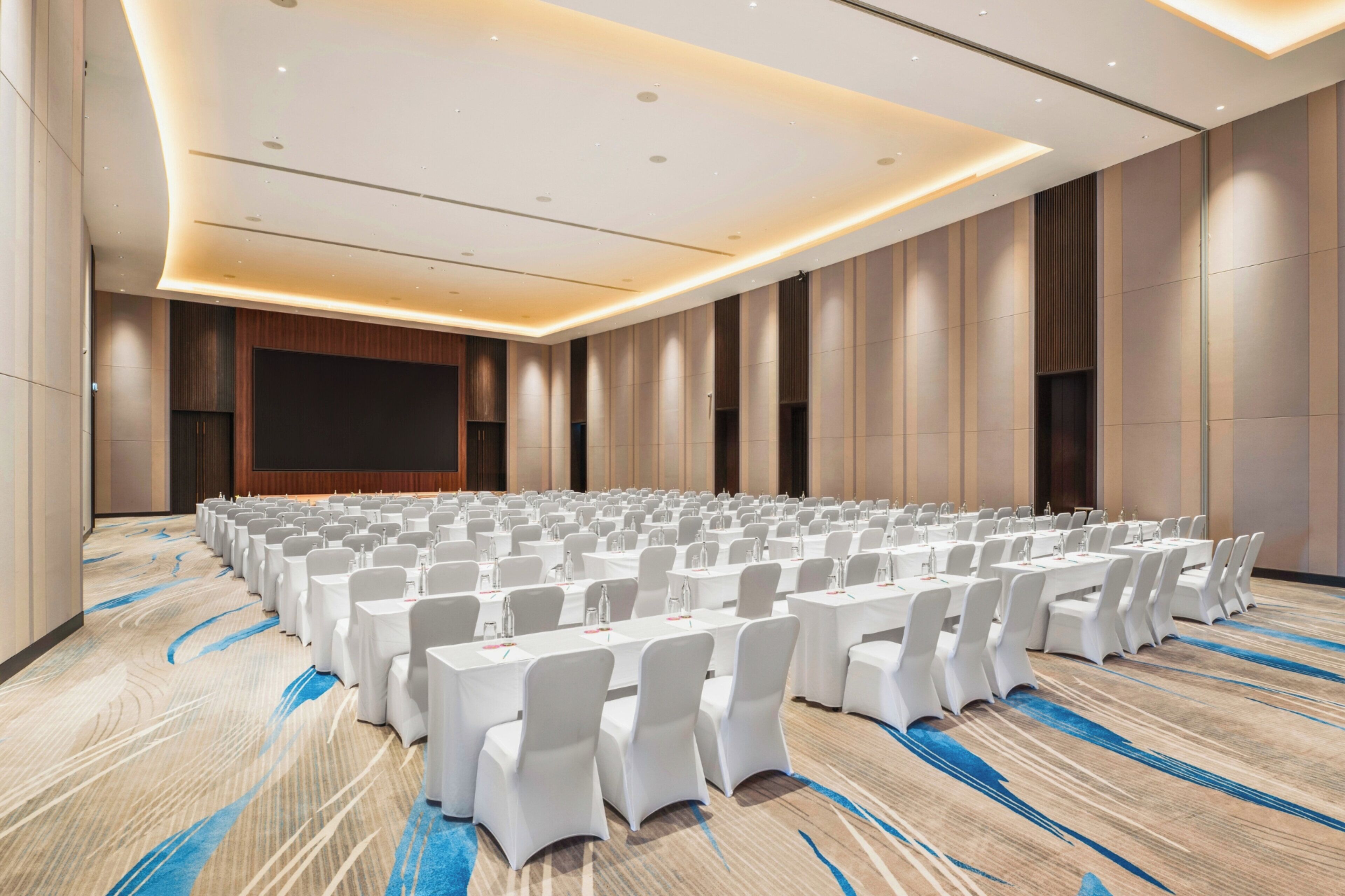 A conference room with rows of tables and chairs, a large screen, and a carpeted floor.