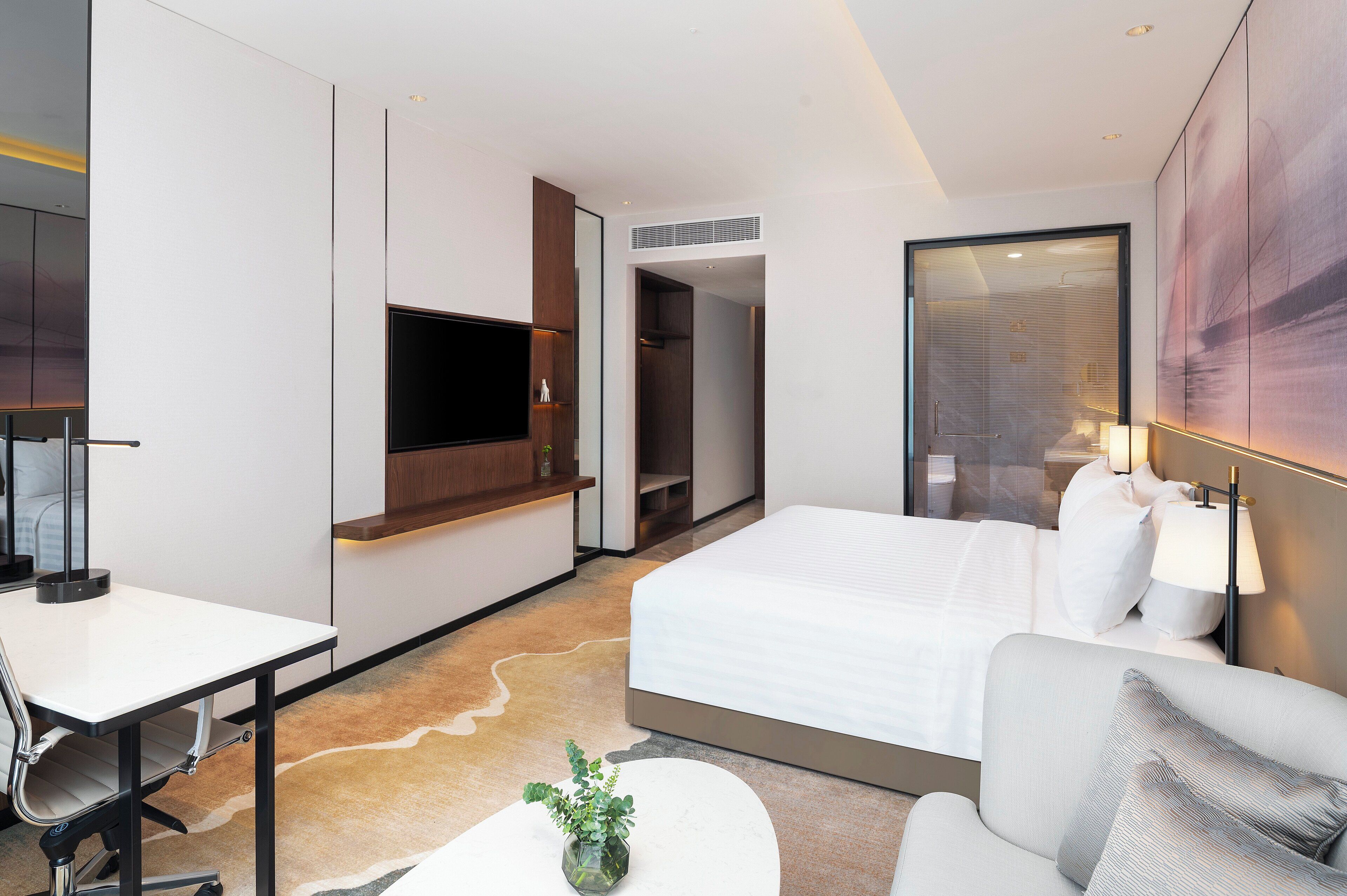 Una habitación de hotel moderna con una cama, un escritorio, una silla, un televisor y un ventanal grande.