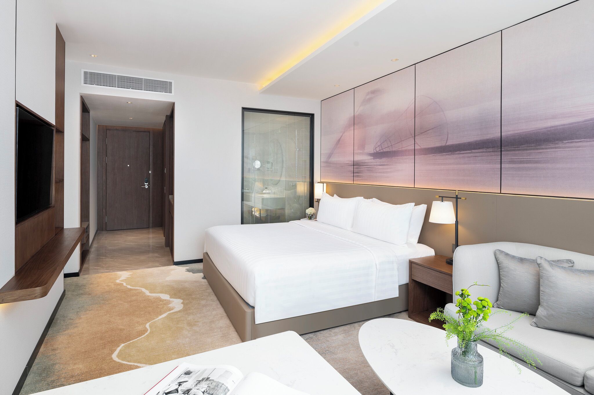 A modern hotel room with a bed, a sofa, a TV, a wall-mounted artwork, and a bedside table with a lamp.