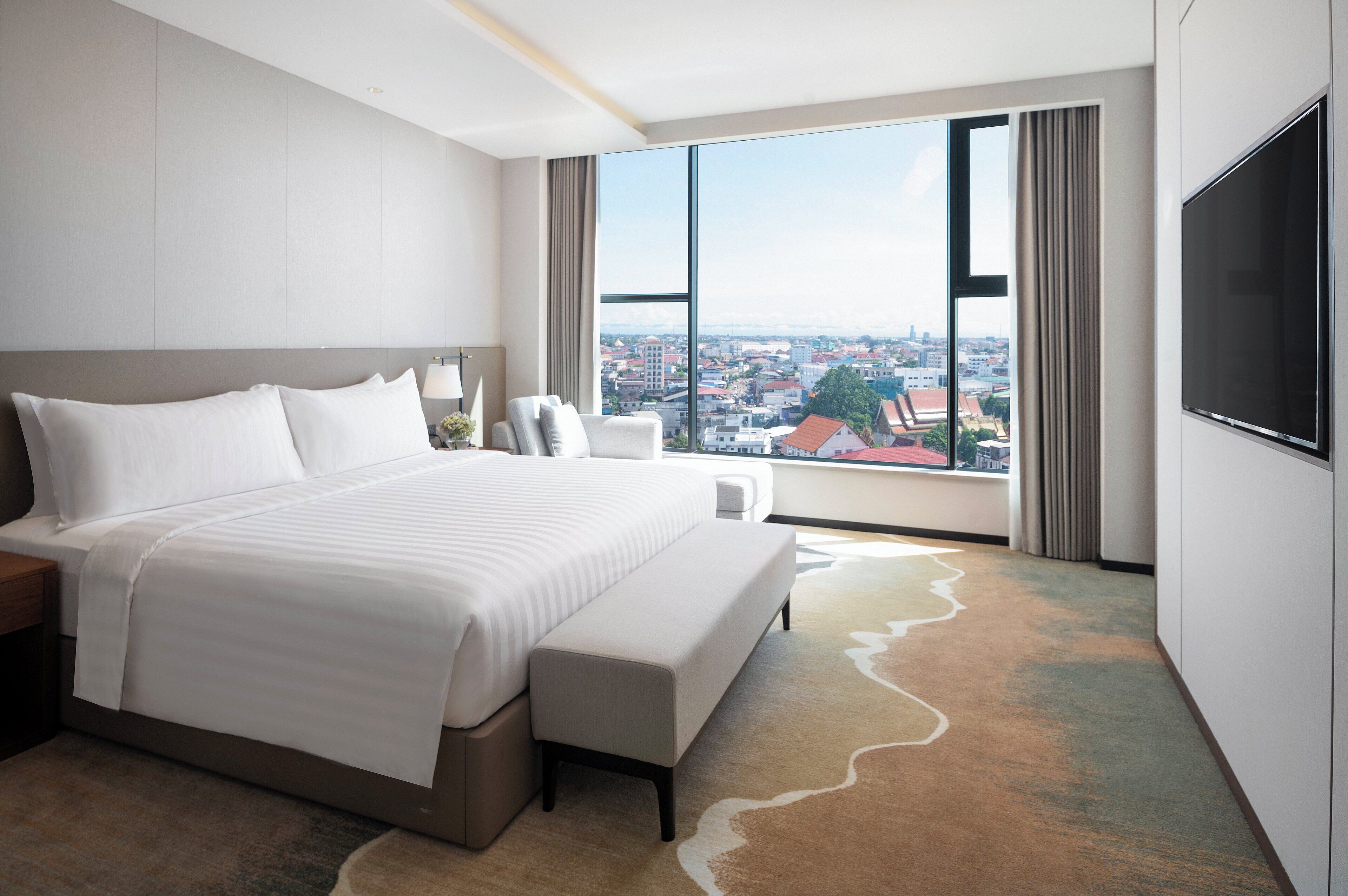 Una habitación de hotel moderna con una cama grande, televisión de pantalla plana, vista a la ciudad y un ventanal amplio.