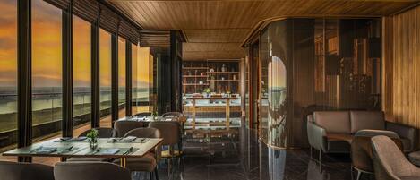 A modern dining area with a view of the landscape through large windows, a wooden ceiling, and a bar area with shelves.