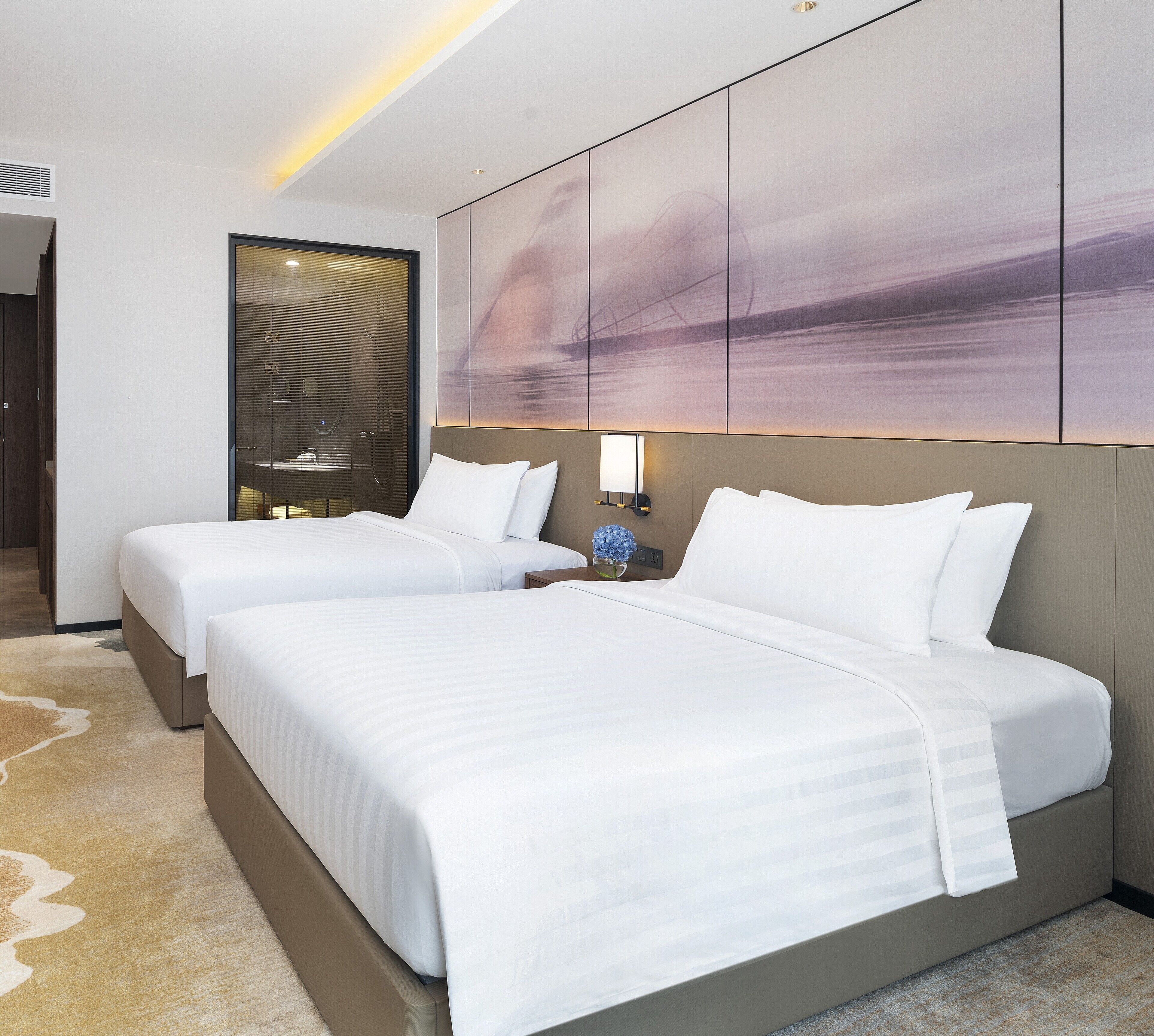 A hotel room with two beds, a large abstract painting on the wall, a nightstand with a lamp, and a view of the city through the window.