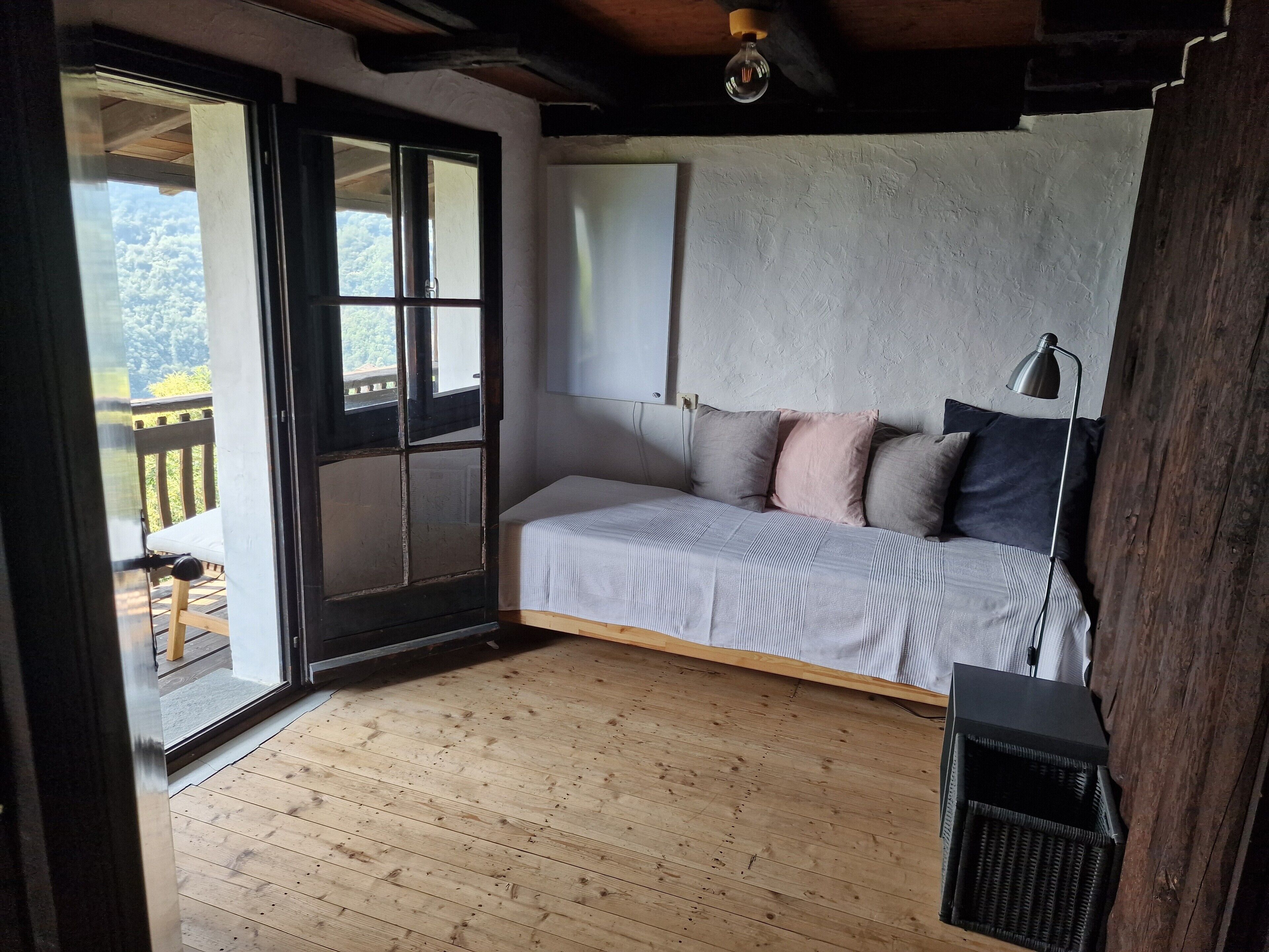 Une pièce avec un plancher en bois, un lit avec des oreillers, un bureau avec une lampe et une fenêtre donnant sur des arbres.