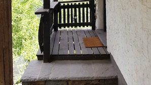 A wooden porch with a stone bench, a view of a forested mountain, and a white wall with a rustic door.