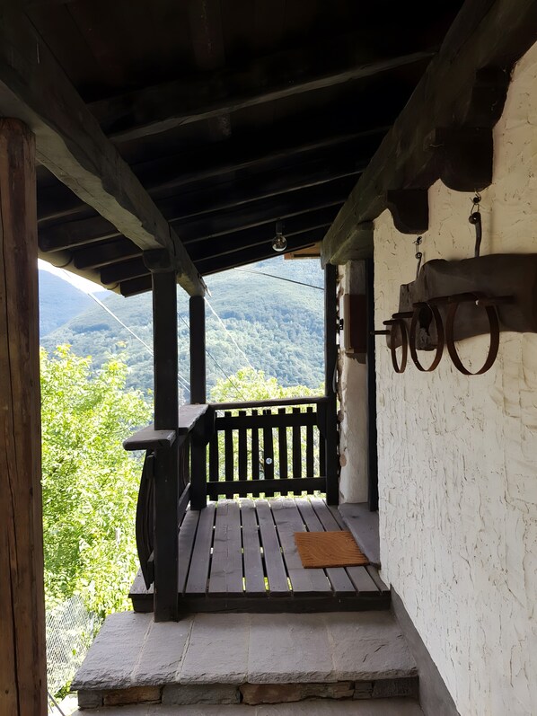 Varanda de madeira com um banco de pedra, vista para uma montanha arborizada e uma parede branca com porta rústica.