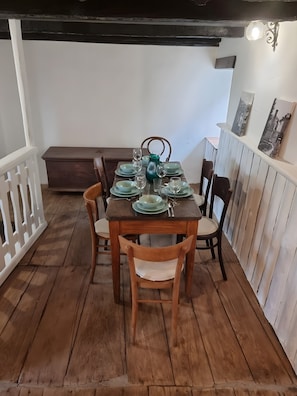 Mesa posta com pratos, copos e um vaso, rodeada por cadeiras, num quarto com chão de madeira e paredes brancas.