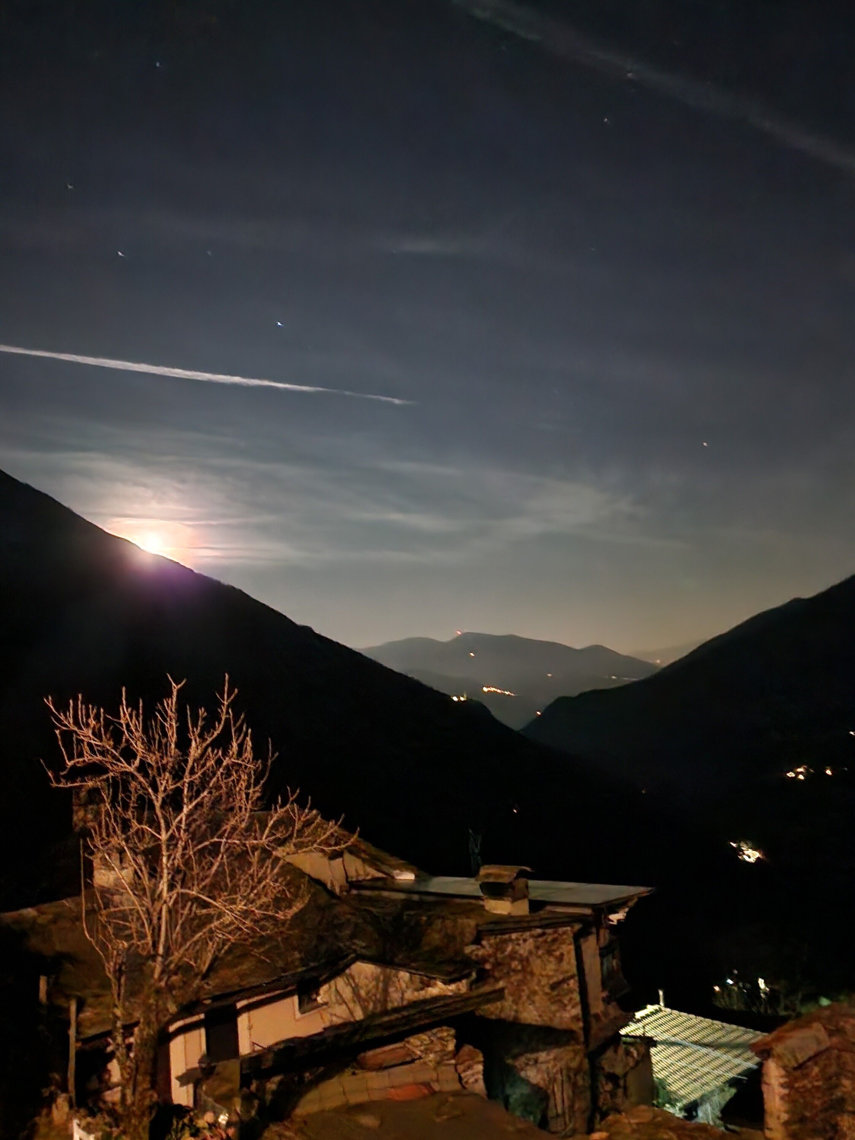 Paisagem montanhosa noturna, com a lua visível, céu limpo e luzes ao longe.