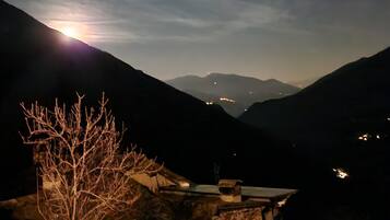Paisagem montanhosa noturna, com a lua visível, céu limpo e luzes ao longe.