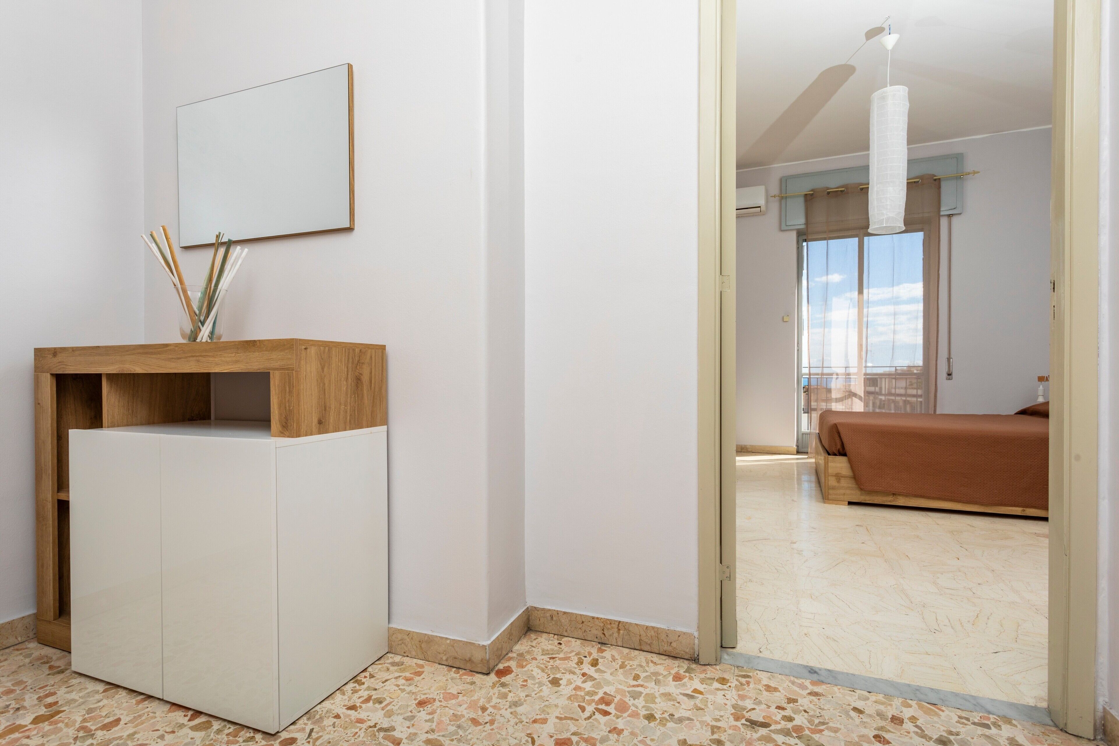Una habitación con un tocador de madera, un espejo, una cama con colcha marrón y vistas a otra habitación por una puerta de vidrio.