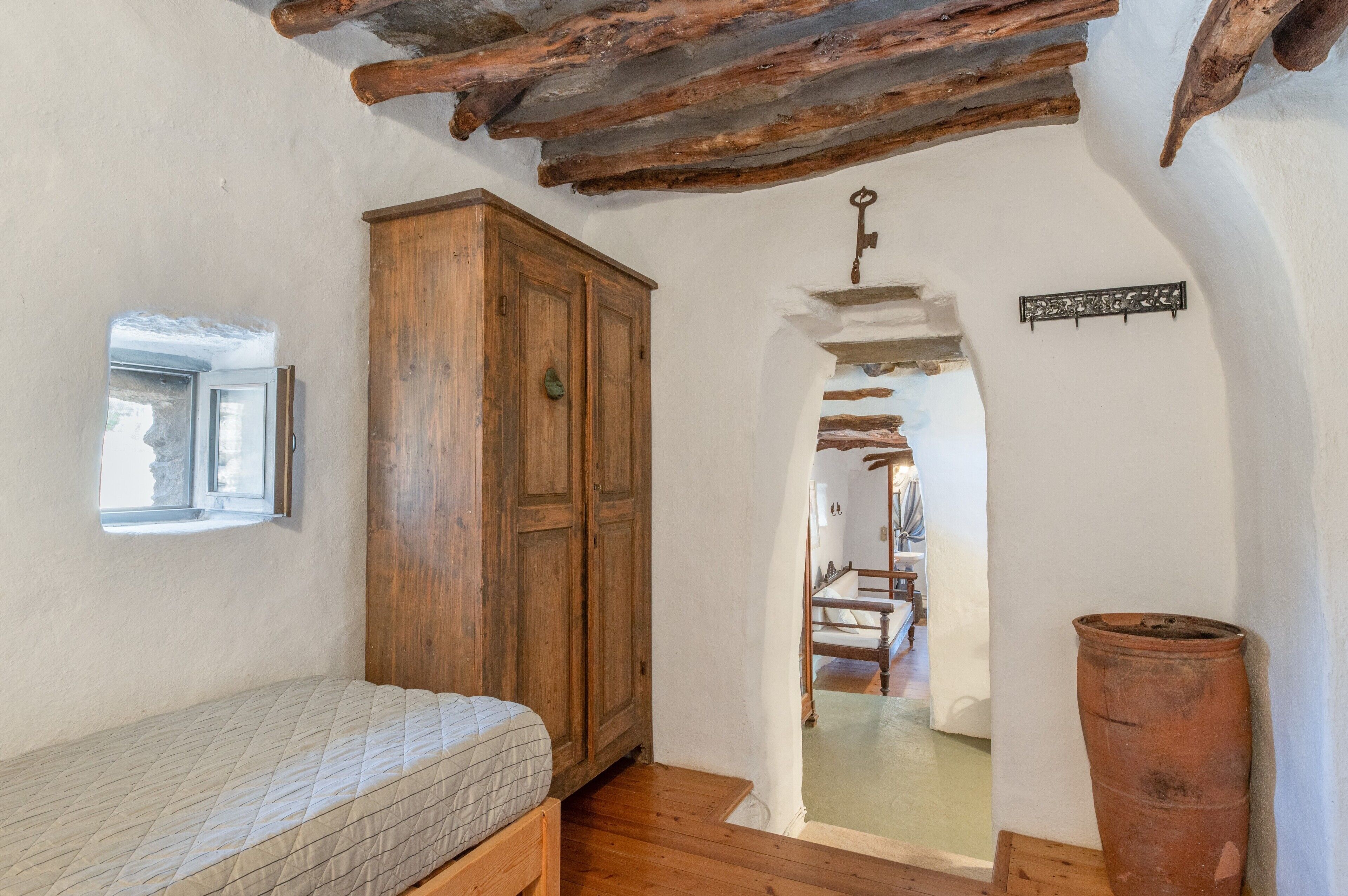 A room with a wooden wardrobe, a mattress, a window, a wooden door, and a rustic ceiling.