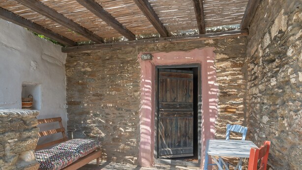 A stone-walled room with a wooden bench, a table with chairs, and a wooden door. - Holiday Home 'Stone House By Anemos' with Private Terrace and Private Garden (Agios Ioannis Tinos)