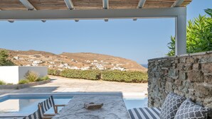 An outdoor seating area with a stone table and chairs, a stone wall, and a view of a hillside with white buildings. - Apartment In A Hotel 'Anemos Suite with Private Pool' with Sea View, Wi-Fi and Air Conditioning (Laouti Tinos)