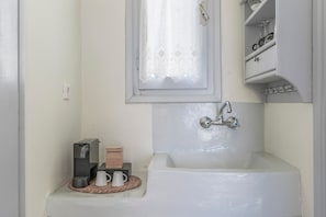 Um banheiro com banheira, uma janela com cortinas de renda, uma prateleira com artigos de higiene pessoal e uma pequena mesa com chaleira e xícaras.
