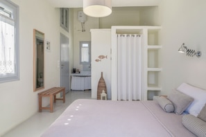 Quarto com cama, um banquinho, guarda-roupa, espelho, prateleira com objetos decorativos e um banheiro ao fundo.