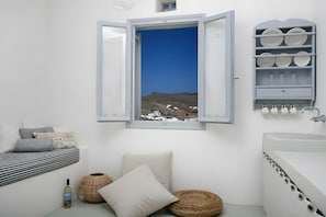 A bathroom with a window view of a white building and landscape, a white sink, and a shelf with plates and glasses.