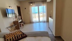 A hotel room with a bed, a chessboard, a TV, and a view of the beach through the window. - Aqua Luna Bay Beachfront Luxury Apartment (Pigianos Kampos)